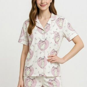 May include: White pajama set with a pink and white heart pattern, featuring a bunny and bow design. The set includes a short-sleeved button-down shirt and matching shorts. The word "PAJAMA" is written in pink at the top.