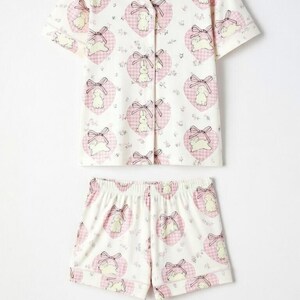 May include: A two-piece pajama set featuring a short-sleeved button-up shirt and matching shorts. The set is white with a repeating pattern of pink hearts, bows, and cartoon bunnies. The shirt has a notched collar and pink gingham trim.