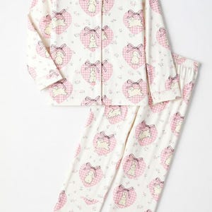 May include: A two-piece pajama set featuring a white background with a repeating pattern of pink gingham hearts, each containing a bunny and bow. The long-sleeved top has a button-down front and the pants have an elastic waistband.