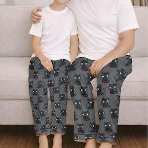 May include: Matching pajama pants featuring a repeating pattern of a black dragon with green eyes and a red tongue on a gray background. The pajama pants are worn by two people, paired with white t-shirts.