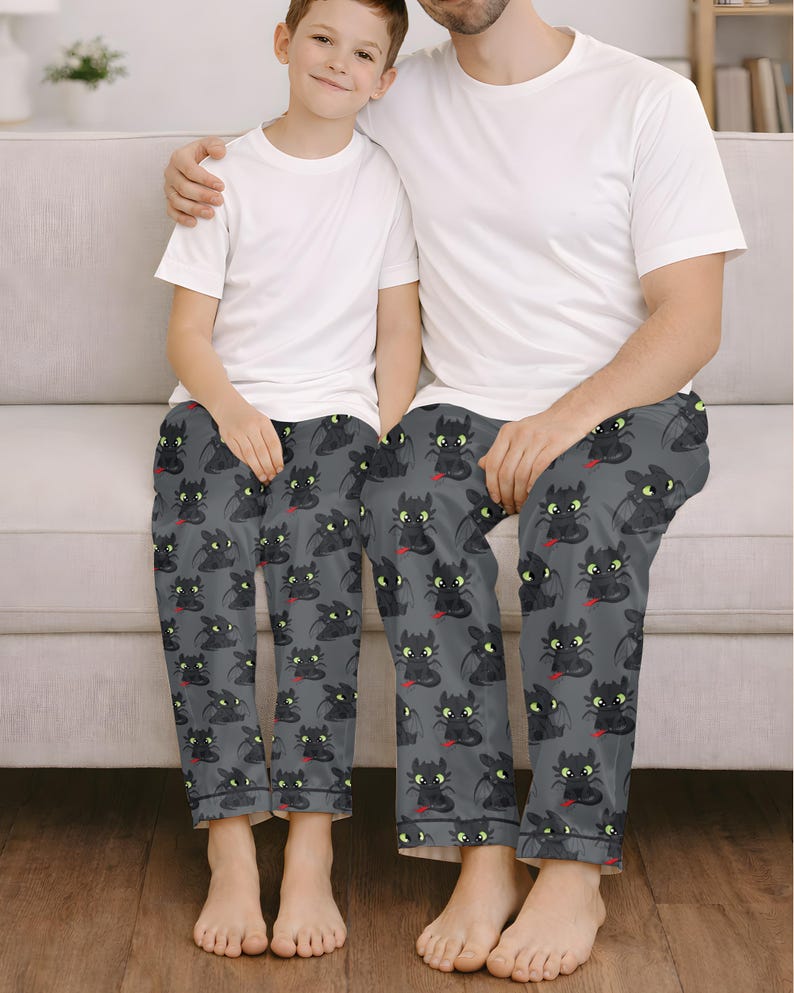 May include: Matching pajama pants featuring a repeating pattern of a black dragon with green eyes and a red tongue on a gray background. The pajama pants are worn by two people, paired with white t-shirts.