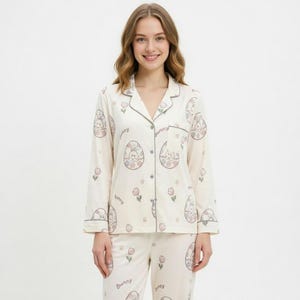 Puede incluir: Conjunto de pijama color crema con un estampado de conejito y huevo de Pascua. La parte superior de manga larga y los pantalones tienen un dise&ntilde;o repetido de conejitos dentro de huevos, flores rosas y la palabra "bunny". El conjunto tiene ribetes grises.