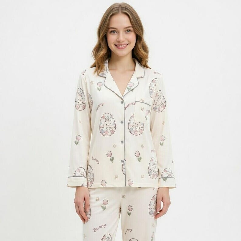 Puede incluir: Conjunto de pijama color crema con un estampado de conejito y huevo de Pascua. La parte superior de manga larga y los pantalones tienen un dise&ntilde;o repetido de conejitos dentro de huevos, flores rosas y la palabra "bunny". El conjunto tiene ribetes grises.
