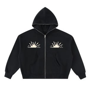 Op de afbeelding: Een zwarte hoodie met ritssluiting en een zilverkleurige sluiting aan de voorzijde. Op de borst staan twee symmetrische, crèmekleurige grafische zonnestraalontwerpen. Het kledingstuk heeft lange mouwen, geribbelde boorden en twee voorzakken.