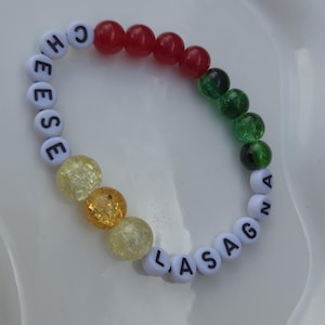 Peut inclure: Un bracelet fait à la main avec les mots "CHEESE LASAGNA" épelés avec des perles blanches. Le bracelet comporte des perles rouges, vertes et jaunes, créant un design coloré et ludique. Pour les amateurs de cuisine.