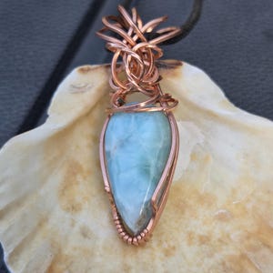 May include: A handcrafted copper wire pendant with a light blue gemstone in a teardrop shape. The gemstone is encased in copper wire, with detailed wirework at the top, suspended from a black cord. The pendant is displayed on a seashell.