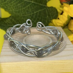 Puede incluir: Un brazalete de alambre plateado con cuentas de piedras preciosas grises. El brazalete hecho a mano tiene un diseño tejido y descansa sobre un pequeño bloque de madera. El fondo incluye hojas verdes y flores amarillas.