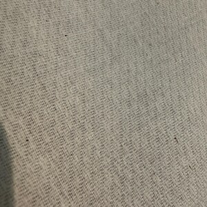 May include: Close-up of a light gray woven fabric with a textured, diagonal pattern. The fabric appears to be made of natural fibers, with a slightly rough and uneven surface. The weave creates a subtle grid-like design.