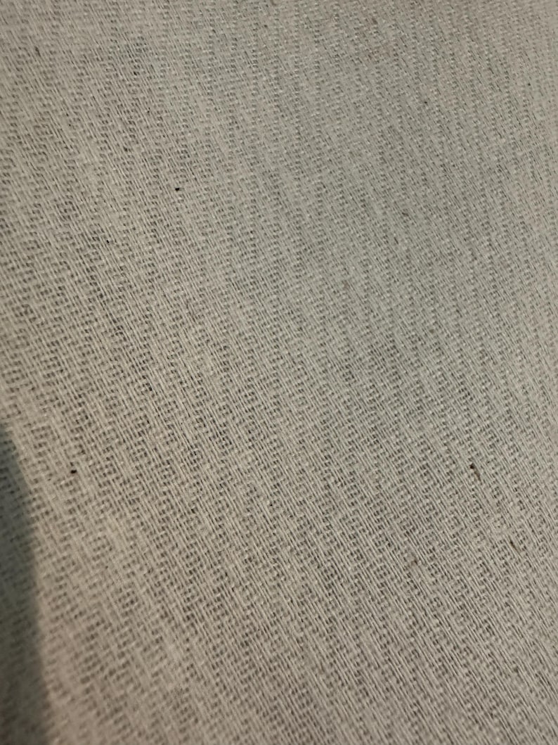 May include: Close-up of a light gray woven fabric with a textured, diagonal pattern. The fabric appears to be made of natural fibers, with a slightly rough and uneven surface. The weave creates a subtle grid-like design.