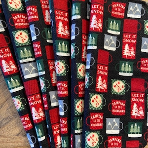 Könnte beinhalten: Ein Stapel schwarzer Stoffservietten mit einem sich wiederholenden Muster aus roten und grünen Tassen, Kompassen und Kiefern. Der Text "LET IT SNOW" und "CAMPING IN THE MOUNTAINS" ist sichtbar.