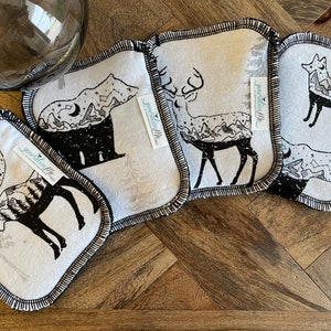 Puede incluir: Juego de cinco posavasos de tela blanca y negra con borde cosido en negro. Cada posavasos presenta una silueta de animal diferente: oso, ciervo, zorro, alce y ciervo. Los posavasos tienen un diseño de montaña y bosque.