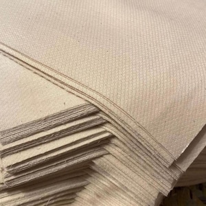 May include: A stack of beige fabric with a subtle diamond pattern. The fabric appears to be folded and layered, with visible edges and a slightly textured surface. The color is a warm, neutral tone, suitable for various textile projects.