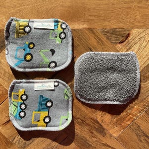 Puede incluir: Tres toallitas grises. Dos tienen un estampado de camiones coloridos y el texto "generation me". La tercera toallita es de color gris liso. Las tres tienen un borde cosido blanco.
