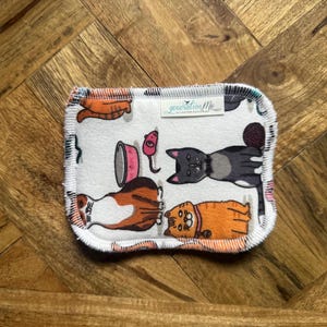 May include: Rectangular white cat-themed cloth with a colorful pattern of cartoon cats, food bowls, and yarn balls. The cloth has a white border and the text "generationMe" in a small label.
