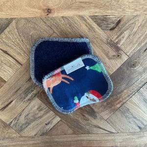 Puede incluir: Dos discos desmaquillantes reutilizables rectangulares de color azul marino. Uno es de color sólido, el otro presenta un estampado navideño con un reno, Papá Noel y un árbol de Navidad. Los bordes están ribeteados con un hilo en contraste.