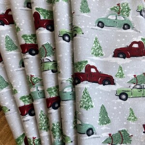 Puede incluir: Conjunto de seis servilletas de algodón grises con un patrón repetido de camiones rojos que transportan árboles de Navidad y coches verdes. Los camiones y los coches conducen por un camino nevado con pinos verdes a ambos lados.