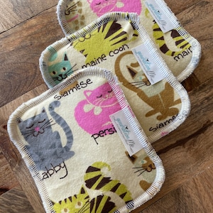 May include: Four beige cat-themed cloths with serged edges are arranged on a wooden surface. Each cloth features a different cat breed illustration, including a gray tabby, a pink Persian, and a brown Maine Coon. The cloths have the text "generation Me" on them.