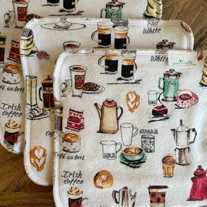 May include: Set of three white cotton dishcloths with a coffee theme. The dishcloths feature illustrations of coffee cups, mugs, pots, and other coffee-related items. The dishcloths have a white background with brown, black, and red accents. The text "Cafe au lait", "Trish Coffee", and "White" is printed on the dishcloths.