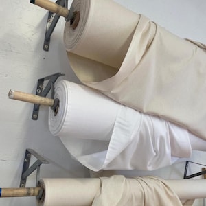 May include: Three rolls of fabric in varying shades of white and beige are mounted on a wall. The rolls are held by metal brackets with wooden dowels. The fabric appears to be cotton or a similar material.