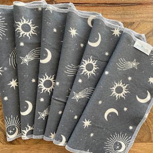 May include: Six gray cotton dishcloths with a white celestial print featuring moons, stars, and suns. The dishcloths are folded and stacked on a wooden surface.