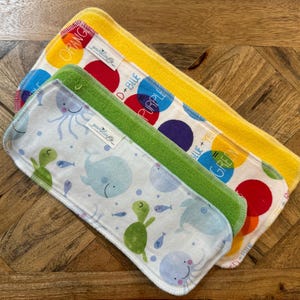 May include: Three rectangular baby cloth wipes. One yellow wipe with the word "ORANGE" and colorful polka dots. One green wipe with sea creatures. One wipe with the words "PURPLE" and "BLUE" with colorful polka dots. All wipes have white trim.