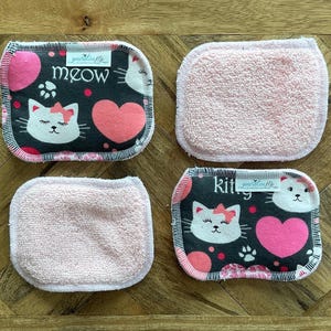May include: Four reusable facial rounds. Two rounds feature a black background with a white cat face, pink hearts, and the word "meow". The other two rounds are solid pink.