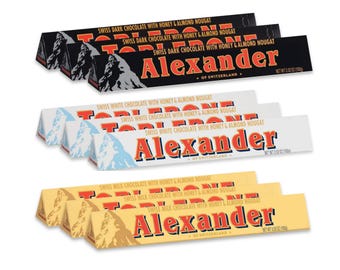 Può includere: Un assortimento di barrette di cioccolato svizzero fondente, bianco e al latte. Ogni barretta presenta il marchio "Alexander" e il testo "Swiss Dark Chocolate with Honey & Almond Nougat", "Swiss White Chocolate with Honey & Almond Nougat", o "Swiss Milk Chocolate with Honey & Almond Nougat".