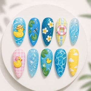 May include: A set of ten painted artificial nails with summer-themed designs. The designs include a yellow duck with a hat, a dragonfly, white flowers, a life preserver, water droplets, a pink grid with a duck, bubbles, a frog, water ripples, and clouds.