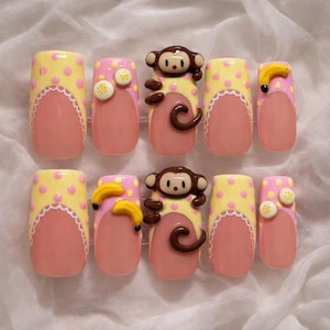 May include: A set of ten press-on nails with a monkey theme. Each nail has a pink base, yellow polka dots, and a white scalloped design. Some nails feature 3D monkey or banana accents.