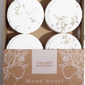 May include: A brown cardboard box containing four round, white shower steamers topped with dried botanical pieces. The box features the text ANCIENT WISDOM, Mood Boost, and SHOWER STEAMERS, along with white line art illustrations of citrus fruits.