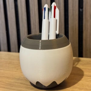 May include: A white and gray pen holder with a rounded shape, holding three white pens with red, blue, and green accents. The pen holder has a unique design with a gray top and a white body.