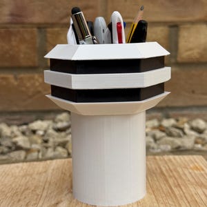 May include: A white and black geometric pen holder with a cylindrical base. The top section has a tiered design with alternating black and white layers. Various pens and pencils are visible inside the holder, including a stylus and a pencil.