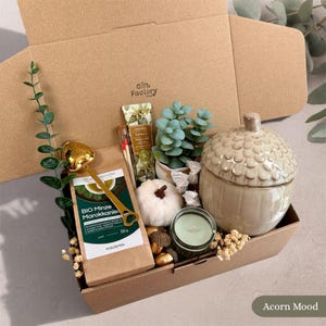 Könnte beinhalten: Eine Geschenkbox mit einer Packung Bio Minze Marokkanisch Tee, einem goldenen herzförmigen Tee-Ei, einer Vanille-Handcreme, Streichhölzern, einer kleinen künstlichen Sukkulente, einem weißen Kürbis, einer hellgrünen Kerze und einem Keramikgefäß in Eichelform.