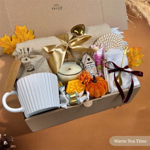 Könnte beinhalten: Eine Geschenkbox mit einer weißen gerippten Tasse, einem Jutebeutel, einer beigen Decke mit goldener Schleife, einer Kerze im Glas, einem kleinen Hausornament, einer Kürbisfigur, einem goldenen Löffel mit dunkelroter Schleife und gelben Blättern.