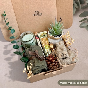 Könnte beinhalten: Eine Geschenkschachtel aus Karton mit einer hellgrünen Kerze im Glas, einer kleinen Sukkulente im Topf, einer Tube Vanille-Handcreme, einem Glasröhrchen mit Streichhölzern, olivgrünen Socken mit braunem Band, einem Tannenzapfen und einem Jutebeutel.