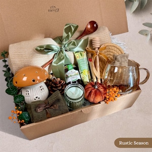 Könnte beinhalten: Eine Geschenkbox mit einer beigen Wohndecke, die mit einem grünen Band gebunden ist, einer Keramik-Pilzlampe, einem Glas Honig, einer Avocado-Handcreme, einem Honiglöffel aus Holz, einer Glastasse, einer kleinen Kerze, einem Stoffkürbis und einem Tannenzapfen.