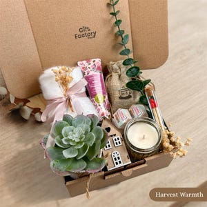 Könnte beinhalten: Eine Geschenkbox mit einem weißen Handtuch mit rosa Schleife, einer floralen Handcremetube, einem Jutebeutel, einem kleinen Holzhaus-Ornament, einer Kerze im Glas, einem Glasröhrchen mit Streichhölzern, einer Sukkulente und Eukalyptuszweigen.