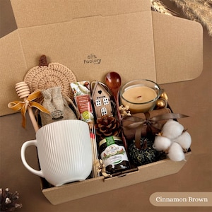 Könnte beinhalten: Eine Geschenkbox mit einer weißen, gerippten Keramiktasse, einem hölzernen Honiglöffel mit goldenem Band, einem gehäkelten Kürbis, einem kleinen Jutebeutel, einem Holzhaus-Ornament, einem Honigglas, einer Kerze im Glas, einem Tannenzapfen und Baumwollzweigen.
