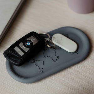 Può includere: Un vassoio ovale grigio con un disegno della mappa del mondo contiene una chiave dell'auto nera con il logo BMW e un portachiavi bianco. Il vassoio è appoggiato su una superficie di legno chiaro. Una tazza rosa è sullo sfondo.