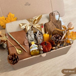Könnte beinhalten: Eine Geschenkbox mit einem braunen Notizbuch, einem goldenen Stift und Löffel, einer beigen Decke mit goldener Schleife, einem Holzbrett, einem Glas Honig, einem kleinen Haus-Ornament, einer gelben Kerze, einem Glasbecher mit Tannenzapfen und Blättern.