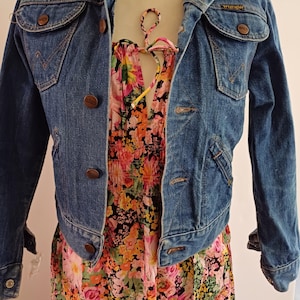 May include: A vintage denim jacket with button fastenings and two chest pockets, worn over a floral print dress. The dress displays a vibrant mix of pink, orange, and yellow flowers. The jacket has the "Wrangler" logo on the pocket.