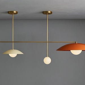 Peut inclure: Un plafonnier moderne à trois lampes. Le luminaire a une finition en laiton avec une barre horizontale et des tiges verticales. Deux lampes ont un abat-jour en demi-dôme, une crème et une orange. La troisième lampe est une sphère blanche.