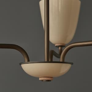 May include: Close-up of a modern chandelier with a cream-colored, bowl-shaped shade and a matching, inverted cone-shaped shade. The fixture has dark bronze-colored arms and a central support, with a neutral gray background.