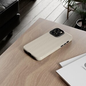 Pu&ograve; includere: Custodia per telefono beige con motivo strutturato e modulo fotocamera nero. La custodia &egrave; appoggiata su una superficie di legno marrone chiaro, con fogli bianchi in primo piano. La custodia &egrave; un accessorio protettivo per telefoni cellulari.