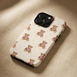Könnte beinhalten: Eine weiße Handyhülle mit einem sich wiederholenden Muster aus braunen Teddybären mit rosa Schleifen und kleinen rosa Herzen. Die Hülle hat ein schwarzes Kameramodul und schwarze Ränder.