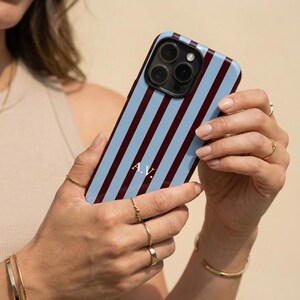 Könnte beinhalten: Eine Handyhülle mit hellblauem Hintergrund und vertikalen weinroten Streifen. Die Hülle hat ein schwarzes Kameramodul und die Initialen "A.V." in Weiß. Die Hülle wird von einer Person mit Goldschmuck gehalten.