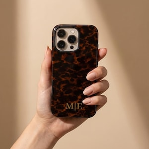 Könnte beinhalten: Eine Handyhülle mit Schildpattmuster in Dunkelbraun und Schwarz. Die Hülle hat eine glänzende Oberfläche und die goldenen Initialen "M|E" unten. Die Handyhülle ist für ein Smartphone mit mehreren Kameralinsen konzipiert.