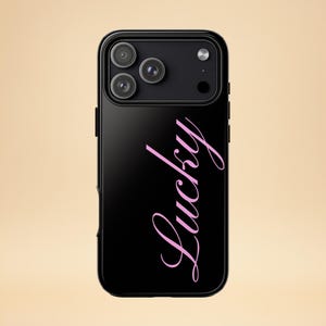 Könnte beinhalten: Schwarze Handyhülle mit glänzendem Finish. Die Hülle hat ein Dreifach-Kamerasystem und das Wort "Lucky" in rosa Schreibschrift. Die Handyhülle wird vor einem hellbeigen Hintergrund gezeigt.