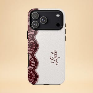 Puede incluir: Funda de teléfono blanca con un diseño de encaje burdeos en el lado izquierdo y los bordes. El nombre "Lale" está escrito en cursiva en el lado derecho. La funda del teléfono tiene una cámara de triple lente.
