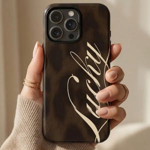 Könnte beinhalten: Dunkelbraune iPhone-Hülle mit dem Wort "Lucky" in weißer Schreibschrift. Die Hülle hat ein gesprenkeltes Muster und einen schwarzen Rand. Die Handyhülle hat drei Kameralinsen.